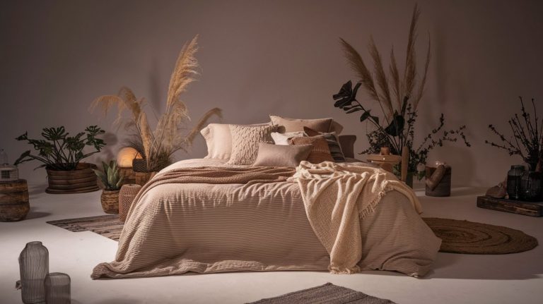 Cozy bedroom setup with a neatly made bed featuring beige linens and an assortment of plush pillows. Dried pampas grass and leafy plants decorate the background, creating a relaxed, natural ambiance in soft lighting.